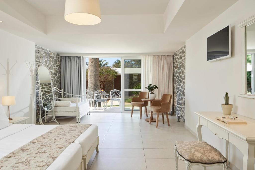 Gaiša un moderna viesnīcas numura iekārtojums ar akmens sienu, divguļamo gultu un ērtu atpūtas zonu. Logi ved uz ārējo terasi, kur redzami palmokoki un dārza augu elementi.