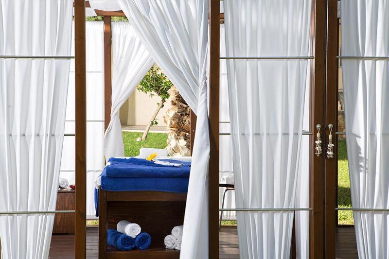 Spa procedūru telpa ar baltām aizkariem un zilu masāžas galdu. Iekārtojums rada mierīgu un relaksējošu atmosfēru.