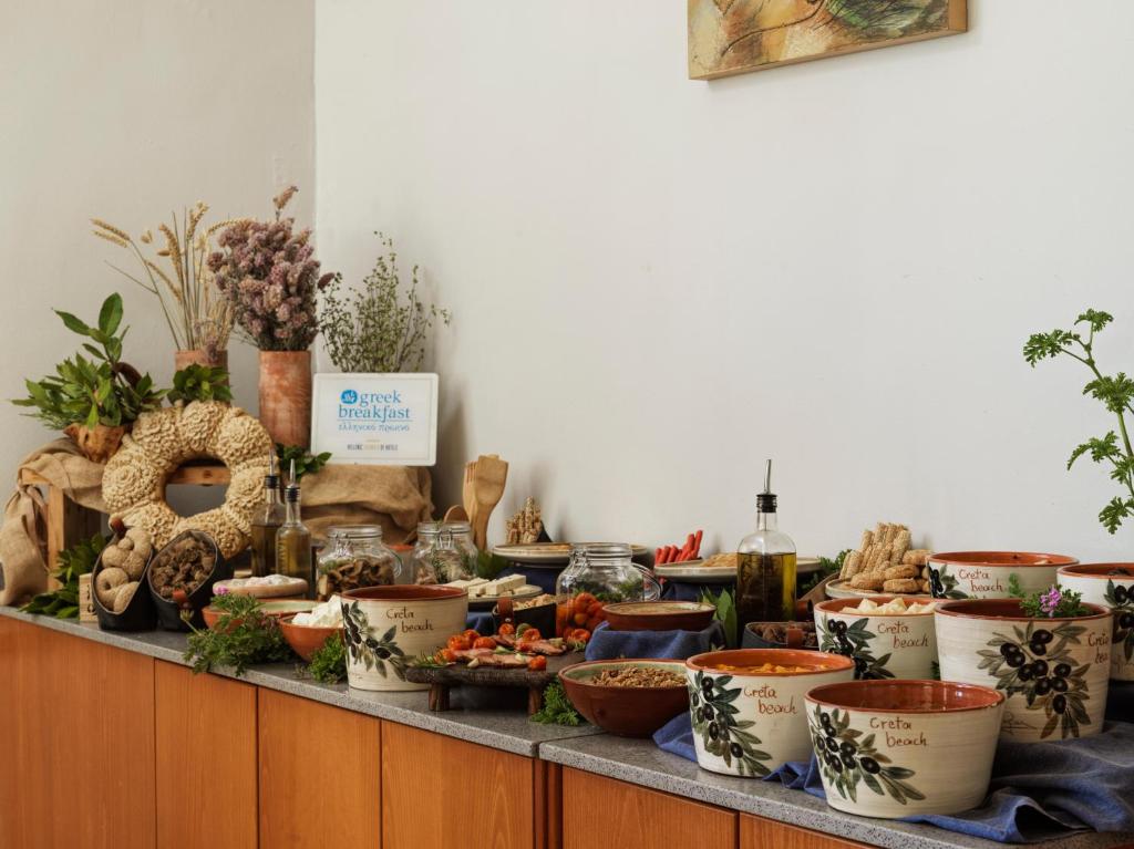 Bagātīgs Grieķu brokastu bufete ar dažādiem ēdieniem un dabīgiem dekoratīviem elementiem, tostarp garšaugiem un olīveļļu. Grozi ar tradicionāliem produktiem un māla trauki piešķir autentisku atmosfēru.