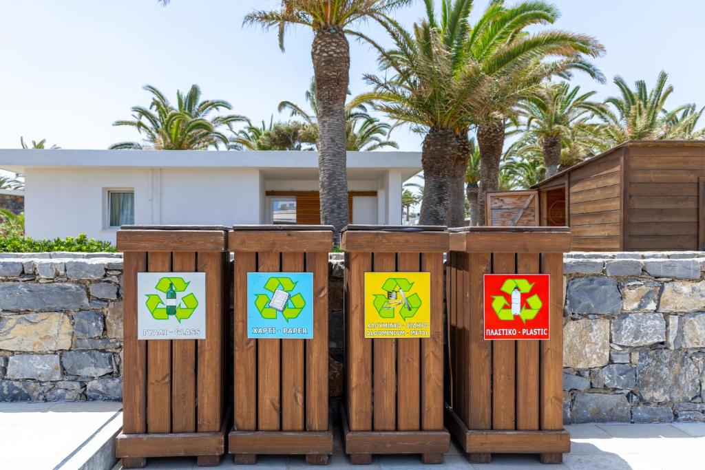 Pārstrādājamie atkritumu konteineri ar skaidriem simboliem stikla, papīra un plastmasas šķirošanai, iestatīti pie modernas ēkas ar palmām fona.