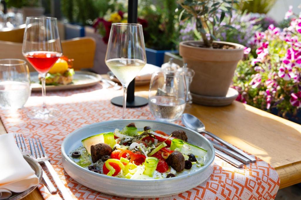 Svaigas un krāsainas salātu porcija ar gaļas bumbiņām, tomātiem, olīvām un dārzeņiem, kas pasniegta pie galda ar vīna glāzēm un ziediem fonā.