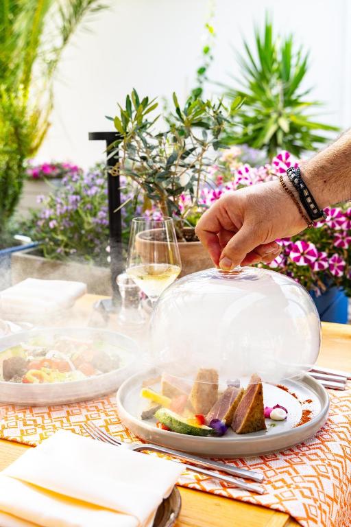 Elegantā pasniegšana ar apklājumu no stikla, atklājot izsmalcinātu ēdienu uz šķīvja, ko papildina krāsaini dārzeņi un ziedi. Fons ar zaļām augu kompozīcijām un ziediem piešķir atmosfēru.