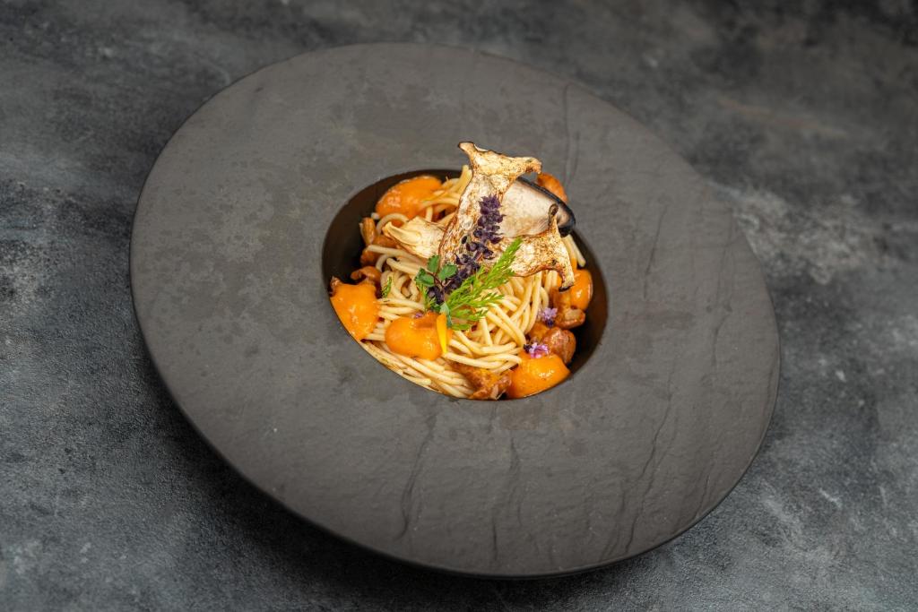 Pasta ēdiens ar sēnēm un garšaugiem, pasniegts melnā traukā uz pelēka fona.