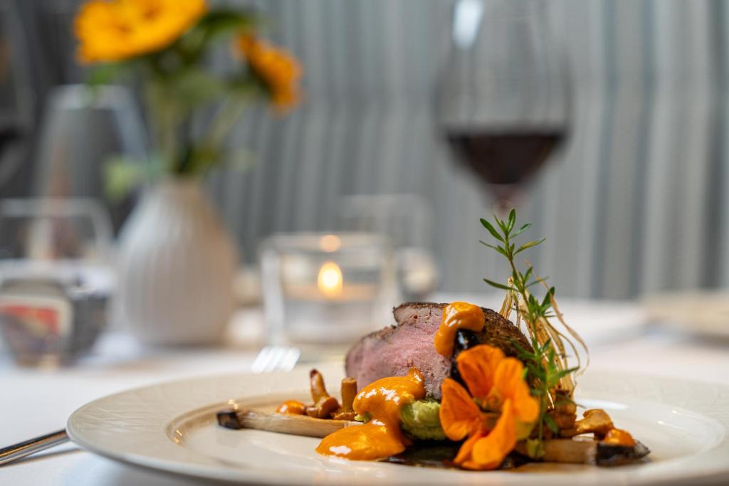 Gurķu un ziedu dekorēts gaļas ēdiens ar mērci, pasniegts elegantā restorānā. Fonā redzama ziedu kompozīcija un vīna glāze.