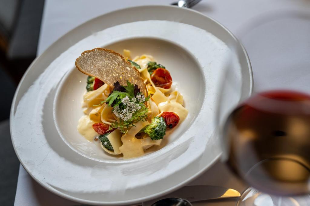 Gards makaronu ēdiens ar krēmīgu mērci, tomātiem un brokoļiem, dekorēts ar garozu un zaļumiem. Fonā redzama glāze sarkanvīna.