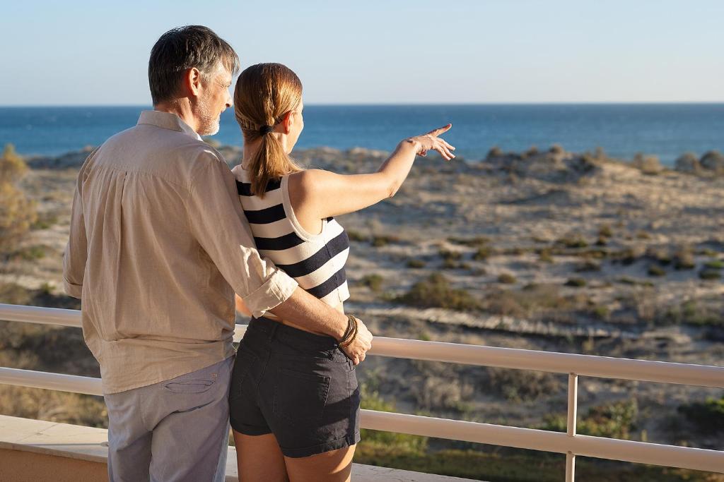 Pāris stāv uz balkona, skatoties uz jūru un norādot uz ainavu. Aiz viņiem redzama saulaina pludmale un zila jūra.
