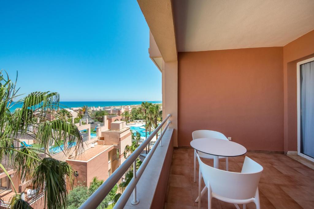 Balkons ar skatu uz jūru, kurā ir balti krēsli un galds, apkārt palmām un peldbaseiniem. Ideāla vieta relaksācijai un atpūtai.