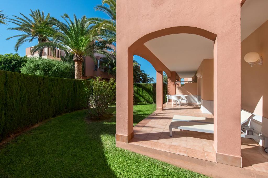 Mūsdienīgs dārza skats ar dzīvokļa terasi, kurā redzami balti sauļošanās krēsli un sulīgas palmas. Terase atrodas blakus zaļai zālei un dzīvžogam, radot relaksējošu atmosfēru.