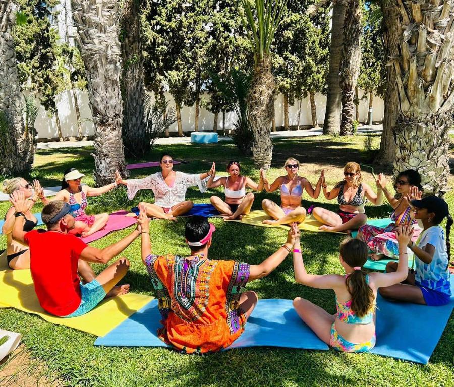 Grupas cilvēku meditācija vai jogas nodarbība zem palmām, sēžot apļveida formācijā uz krāsainām paklājiem.