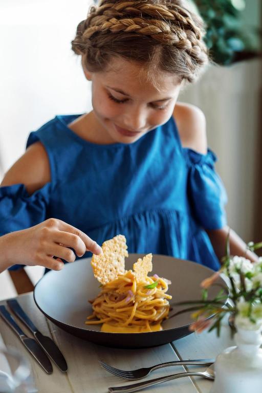 Bērns ar pīnēm, tērpies zila blūze, baudot makaronus ar kraukšķīgu uzkodu, sēžot pie galda.
