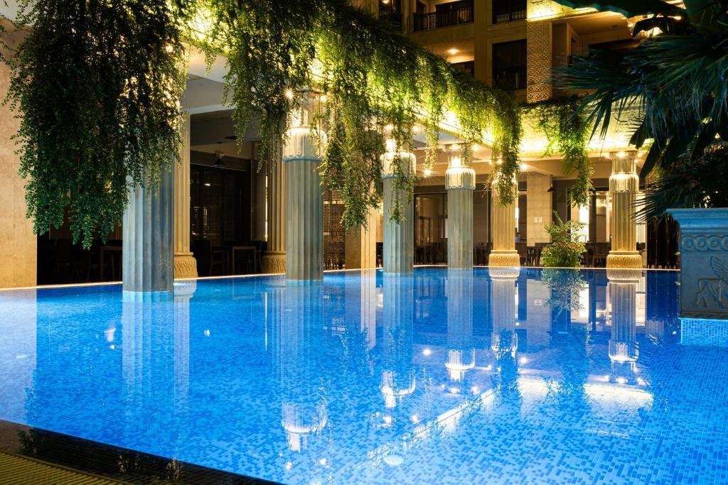 Luksusīgs iekštelpu baseins ar dzidrām ūdens virsmām un greznām kolonnām, apgaismots ar siltām lampām un ieskauts zaļām vīnogulājām.