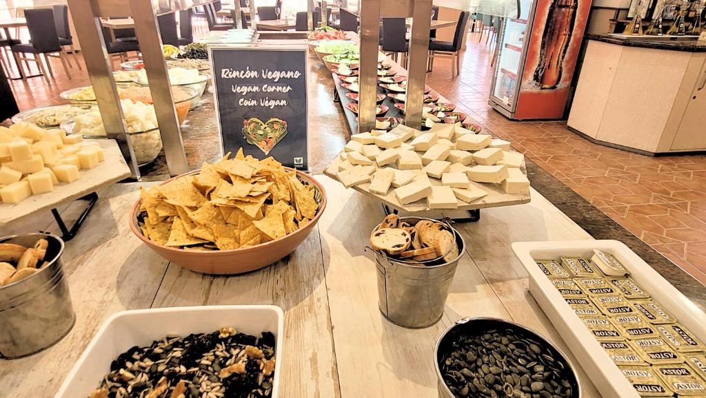 Veganās uzkodas bufete ar dažādiem dārzeņiem, čipsiem, tofu un grauzdiņiem. Uz galda redzama zīme "Rincón Vegano", norādot uz vegānisko stūri.