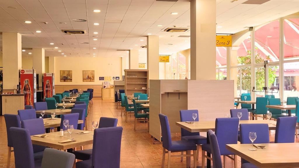 Gaiša un plaša restorāna telpa ar galdiem un krēsliem, kas izkārtoti dažādās zonās. Logi nodrošina dabīgu apgaismojumu, un interjers ir moderns ar zilām un zaļām krāsām.