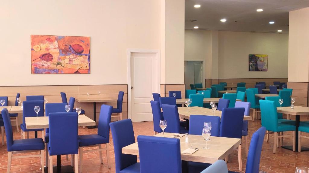 Mūsdienīgs restorāna interjers ar krāsainām zilu un zaļu krēslu grupām un galdiem, papildināts ar mākslas darbu uz sienas.