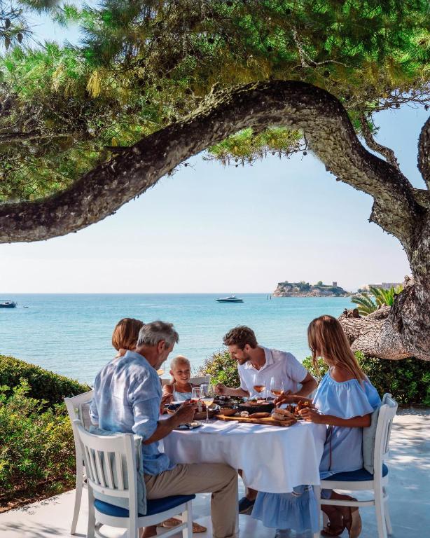Ģimenes vakariņas pie jūras, baudot ēdienus un dzērienus zem koka, ar skatu uz ūdeni un laivām. Atmosfēra ir mierīga un svinīga.