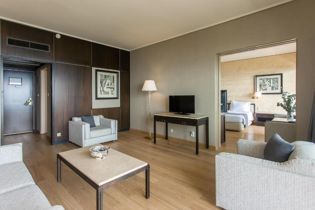 Mūsdienīga viesnīcas numura telpa ar gaišu interjeru, ērtiem dīvāniem un televizoru. Telpa ir dekorēta ar mākslas darbiem un koka grīdu, radot patīkamu atmosfēru.