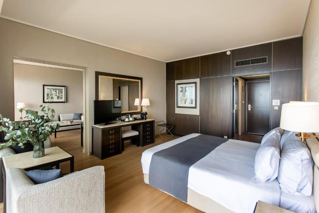 Mūsdienīgs viesnīcas numurs ar elegantām mēbelēm, gultu ar baltām un pelēkām gultasveļām, televizoru un darba galdu. Numurā ir arī atpūtas zona ar dīvānu un dekoratīviem augiem.