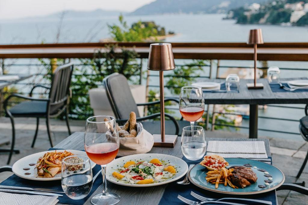 Elegants restorāna galdiņš ar skatu uz jūru, uz kura redzami trīs dažādi ēdieni un dzērieni, radot patīkamu atmosfēru.