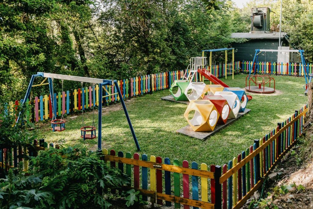 Krāsains bērnu rotaļu laukums ar šūpolēm, slaidu un spēļu elementiem, norobežots ar krāsainu žogu. Zaļa zāle un apkārt esoši koki rada patīkamu vidi bērnu spēlēm.