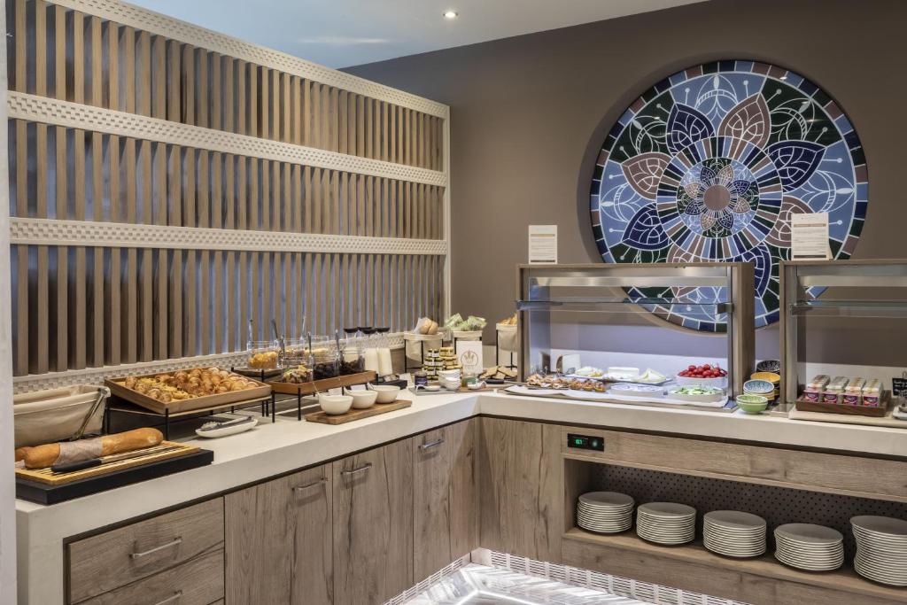 Brokastu zona ar dažādiem ēdieniem, tostarp maizēm un saldumiem, ko papildina eleganta stikla mozaīka uz sienas.
