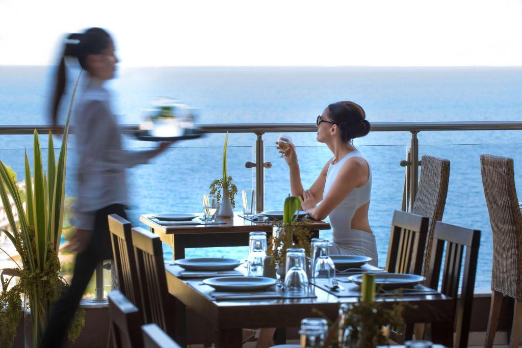 Sieviete bauda dzērienu pie galda Blue Bay Resort Crete terasē ar skatu uz jūru.
