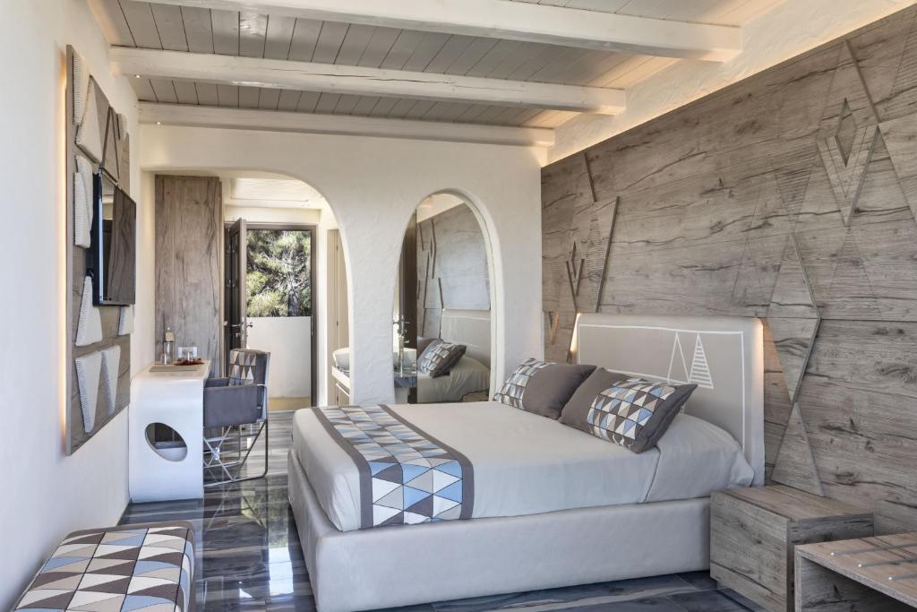 Mūsdienīgs guļamistabas interjers ar koka akcentiem un elegantu apgaismojumu, kas rada mierīgu atmosfēru. Gultā ir dekoratīvi spilveni, un telpā ir pieejama rakstāmgalds un televizors.