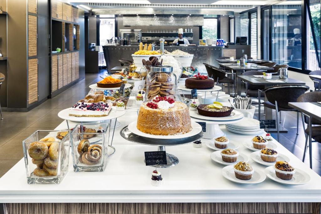 Brokastu bufete ar dažādiem desertiem, tostarp kūkām, maizēm un cepumiem, pie elegantas galda noformējuma. Fons ar virtuvē strādājošu šefpavāru un mūsdienīgu interjeru.