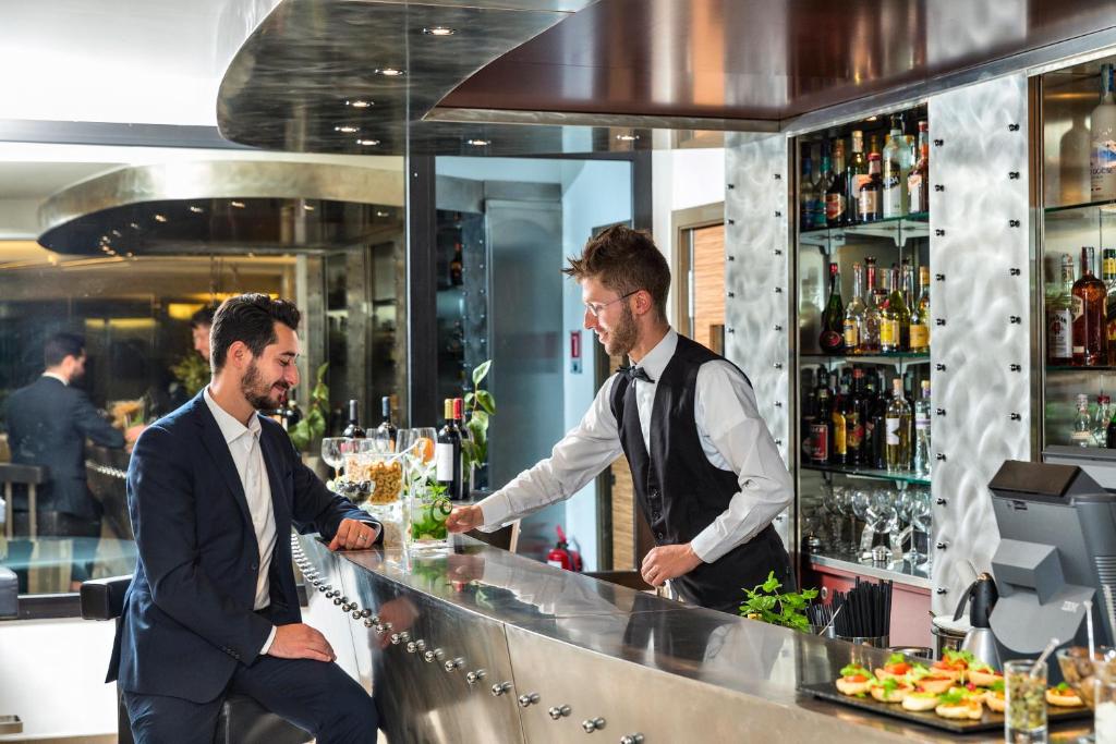 Mūsdienīgs bārs ar viesmīli un apmeklētāju, kur abi sarunājas pie stilīgas, spīdīgas bāra letes. Fons iekļauj plašu alkohola un dzērienu izvēli.