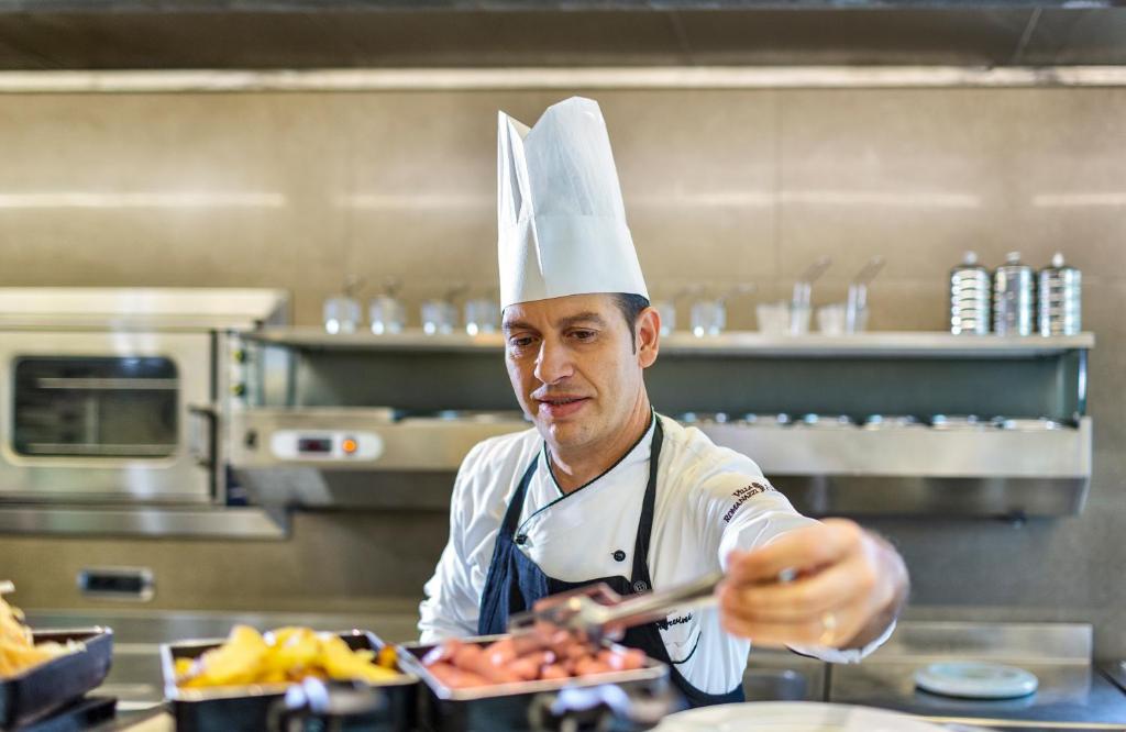 Šefpavārs, tērpies baltā cepurē un uniformā, pasniedz ēdienus virtuvē, izmantojot knaibles. Fons ir moderns un profesionāls.
