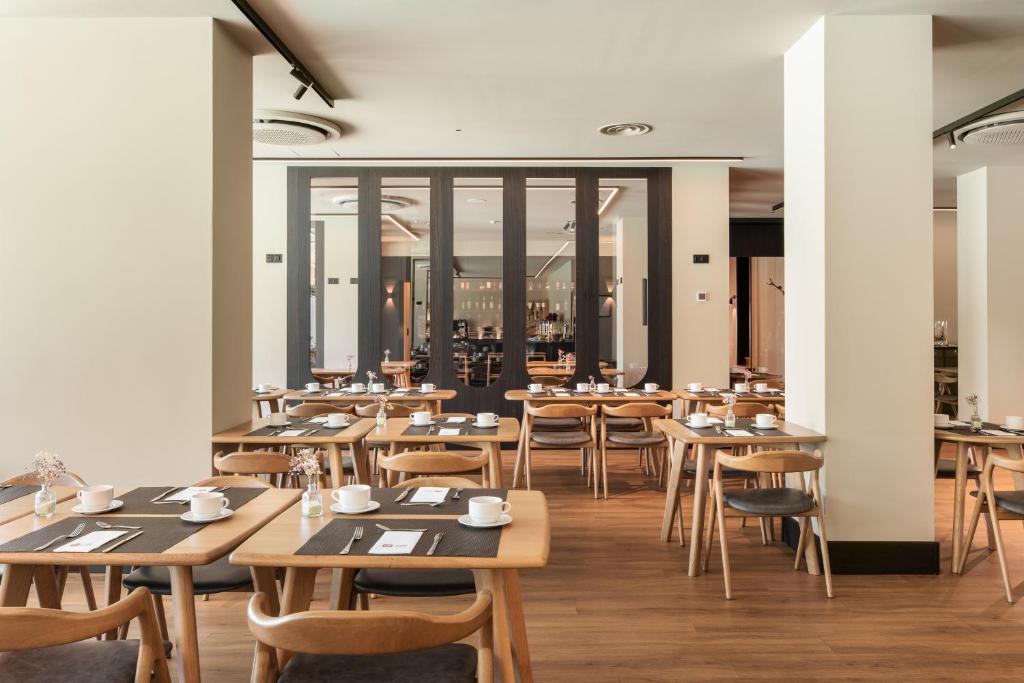 Mūsdienīgs restorāna iekšskats ar koka galdiem un krēsliem, kā arī eleganti sakārtotām galdu klāšanas detaļām. Telpa ir gaiša un plaša, radot patīkamu atmosfēru.