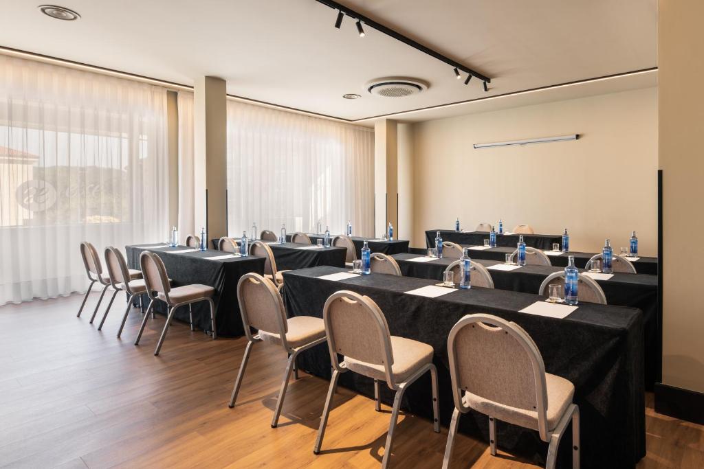 Konferenču telpa ar melniem galdiem, uz kuriem novietoti ūdens pudeles un piezīmju lapas, apgaismota ar dabiskiem gaismas avotiem no logiem.