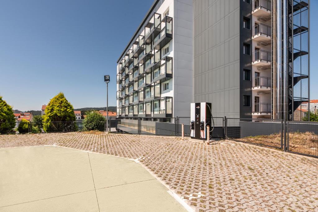 Mūsdienīga daudzdzīvokļu ēka ar pārklātu laukumu un elektrisko automobiļu uzlādes staciju. Apkārtne ir zaļa, ar koku grupām un skaidru zilu debesu fonu.