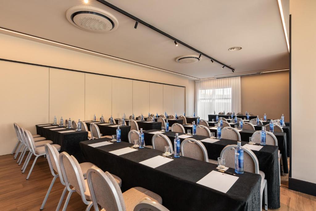 Konferences telpa ar melniem galdautiem, uz kuriem novietotas ūdens pudeles un papīra lapas, ar krēsliem gar abām pusēm. Gaismas no logiem un moderns apgaismojums rada profesionālu atmosfēru.