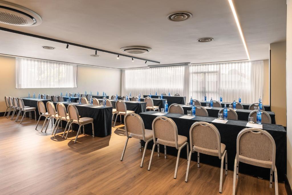 Konferenču telpa ar galdiem un krēsliem, kas sakārtoti divās rindās, katrā ir ūdens pudeles un piezīmju lapas. Telpa ir apgaismota ar dabiskiem gaismas avotiem un modernu apgaismojumu.