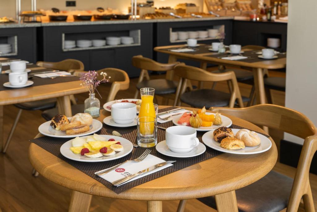 Brokastu galds ar dažādām uzkodām, augļiem un dzērieniem, iekārtots modernā restorānā. Galdā redzami maizītes, augļi un kafija, radot patīkamu un viesmīlīgu atmosfēru.