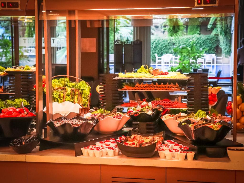 Bagātīgs salātu bārs ar dažādiem dārzeņiem, augļiem un garnējumiem, kas izkārtoti elegantās traukos. Fons rāda viesnīcas interjeru ar zaļām ainavām.