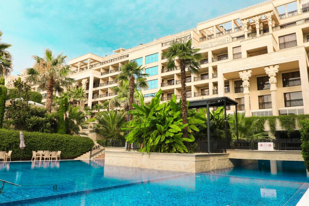 Greznas viesnīcas komplekss ar palmām un baseinu, kas atspoguļo debesu zilo krāsu. Apkārt ir zaļi augi un ainavs.