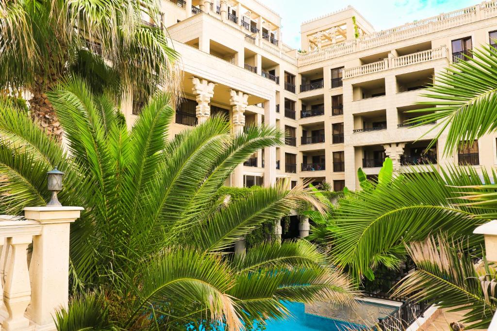 Zaļš tropu ainava ar palmām un viesnīcu kompleksu, kas ieskauj peldbaseinu. Atpūtas vieta, kas piedāvā relaksāciju un komfortu.