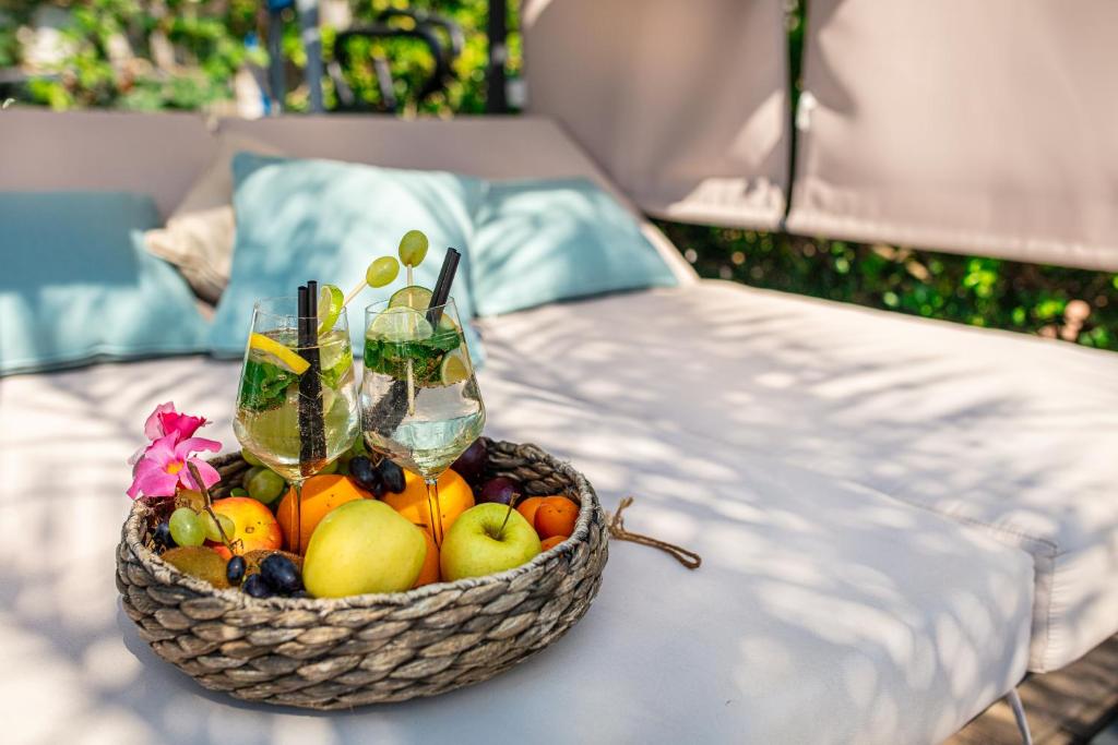 Divas dzirkstošas dzērienu glāzes ar augļu rotājumiem un grozs ar svaigiem augļiem, kas novietots uz atpūtas gultas zemē.