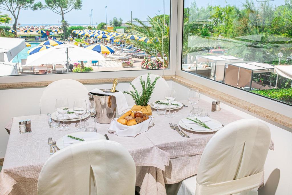 Elegants restorāna galds ar skatu uz baseinu un pludmali, dekorēts ar traukiem un maizītēm. Apkārt ir palmām un saulessargiem, radot relaksējošu atmosfēru.