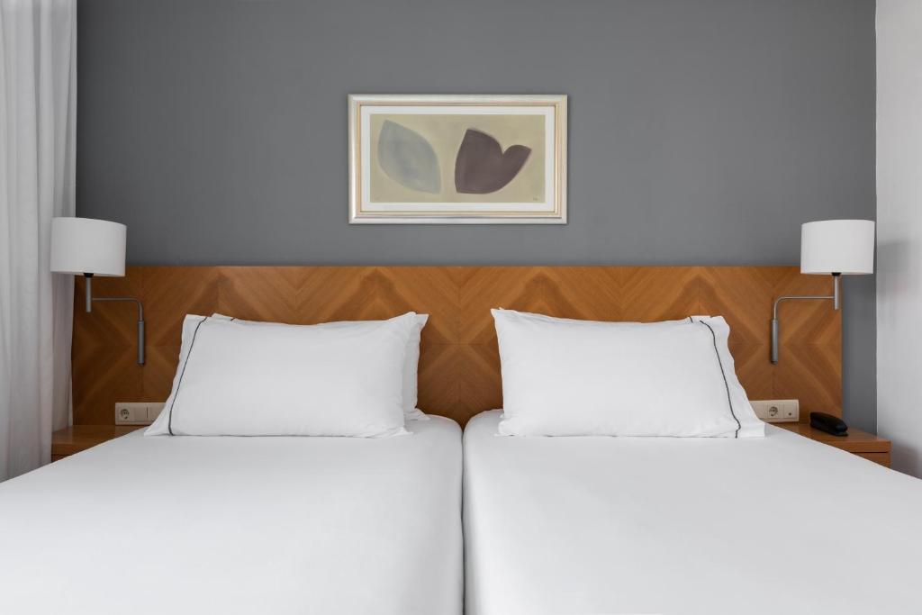 Mūsdienīgs guļamistabas interjers ar divām gultām, koka galvgultu un minimālistisku dekoru uz pelēka fona. Gaismas avoti un mākslas darbs pie sienas piešķir telpai siltumu.