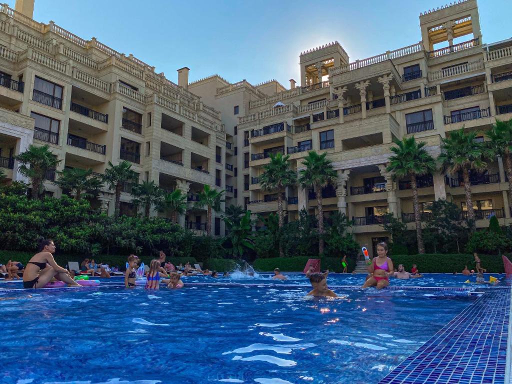 Vasaras brīvdienu ainava pie baseina, kur bērni un pieaugušie izbauda ūdens izpriecas, bet fons veido grezns viesnīcas komplekss ar palmām un saulainu debesu.