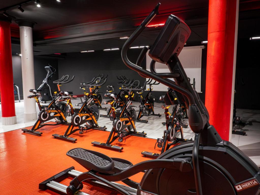 Sporta zāles iekārtojums ar velosipēdu trenažieriem un eliptiskajiem trenažieriem uz sarkanas grīdas. Telpa ir moderni iekārtota ar melniem sienām un spilgtiem elementiem.