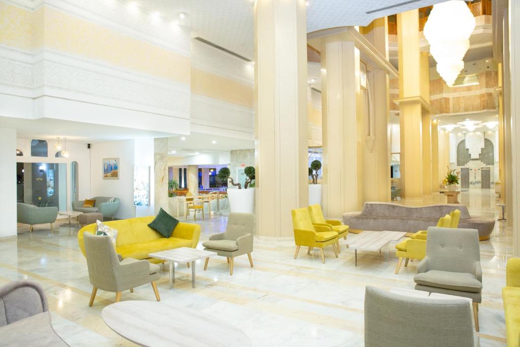 Mūsdienīgs viesnīcas interjers ar gaišām krāsām un elegantu dizainu, kurā redzami komfortabli dīvāni un galdi. Plašais telpas plānojums rada viesmīlīgu atmosfēru.