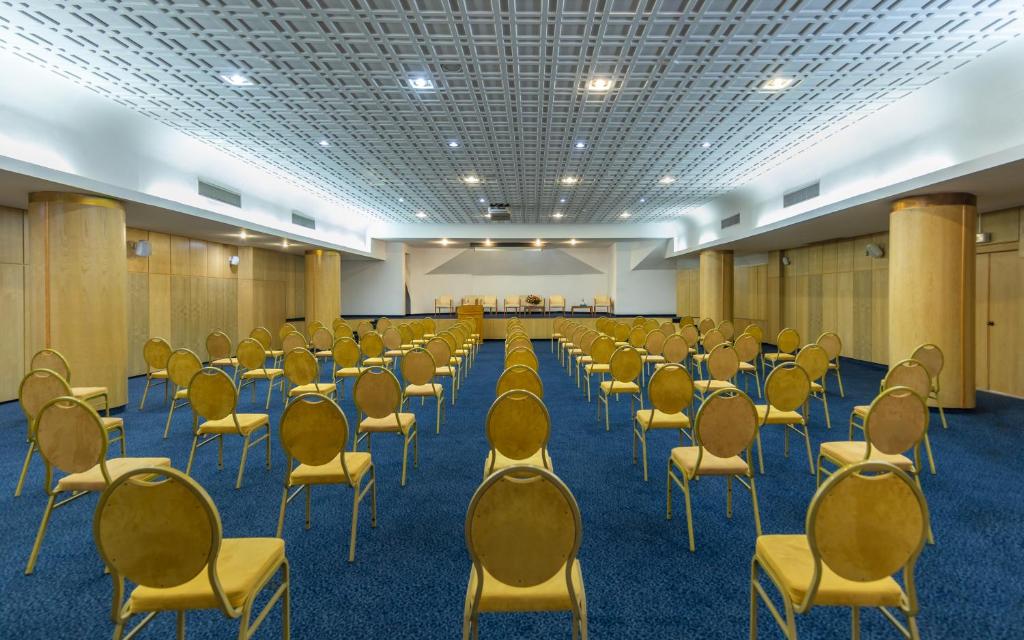 Konferenču zāle ar rindām no dzeltenām krēsliem un gaišu, mūsdienīgu interjeru. Zāles aizmugurē redzama platforma ar galdiem.