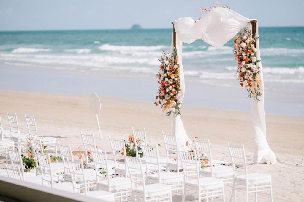 Kāzu ceremonijas vieta pludmalē ar baltām krēsliem un dekoratīvām ziedu kompozīcijām zem svinīgā arka.