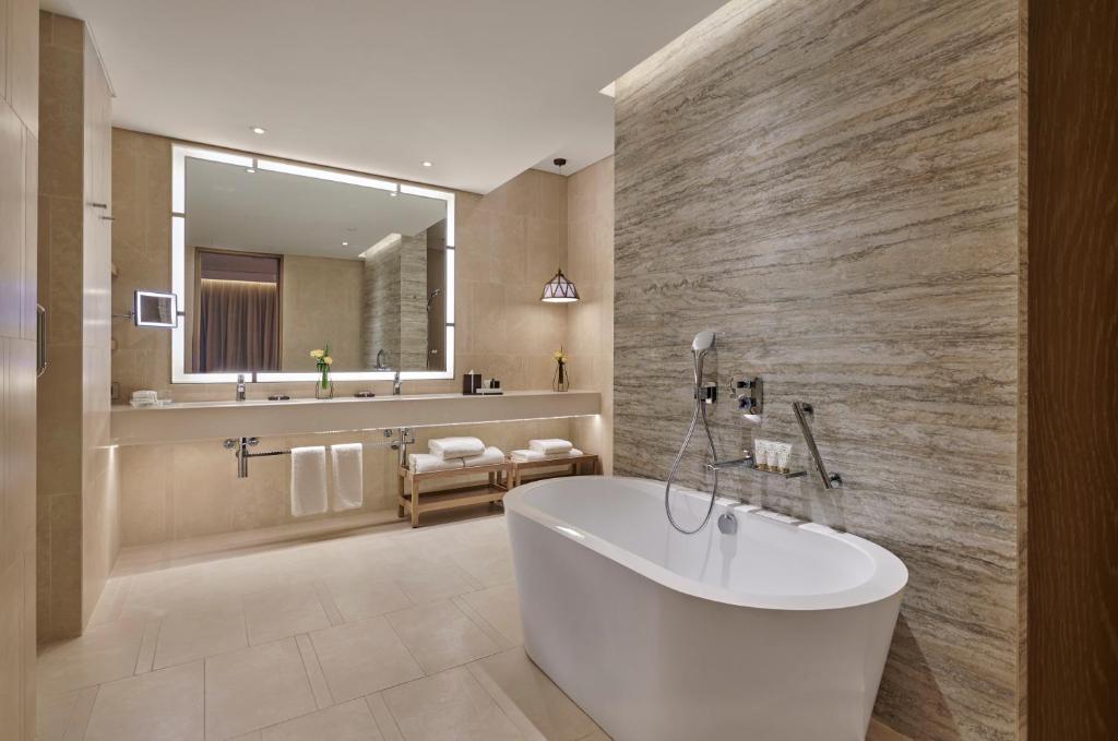 Mūsdienīgs vannas istabas interjers ar brīvdabas vannu, elegantu spoguli un dabīgām akmens sienām. Svaiga zieda klātbūtne pie izlietnes piešķir telpai dzīvīgumu.