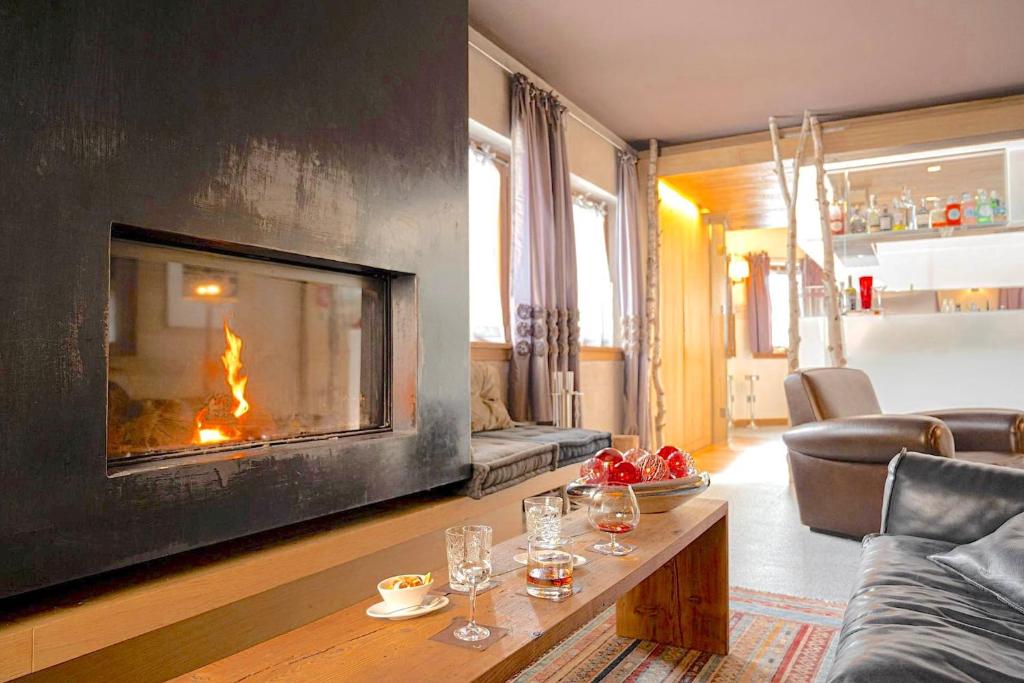 Mūsdienīgs dzīvojamās istabas interjers ar centrālo kamīnu un ērtām mēbelēm, kas rada siltu un viesmīlīgu atmosfēru. Uz galda novietoti dzērieni un uzkodas, papildinot relaksējošo vidi.