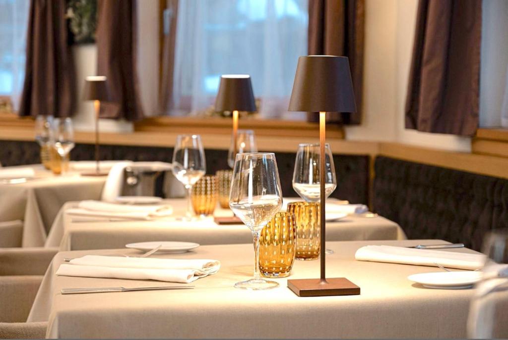 Eleganta restorāna interjers ar galdiem, kas klāti ar baltām galdautām, stikla traukiem un moderna apgaismojuma lampām. Siltas krāsas un mājīga atmosfēra veido aicinošu vidi.