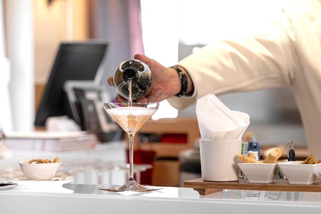 Bārmenis ielej kokteili martini glāzē, kamēr blakus ir uzkodas un salvetes. Eleganta bāra atmosfēra ar modernu interjeru.